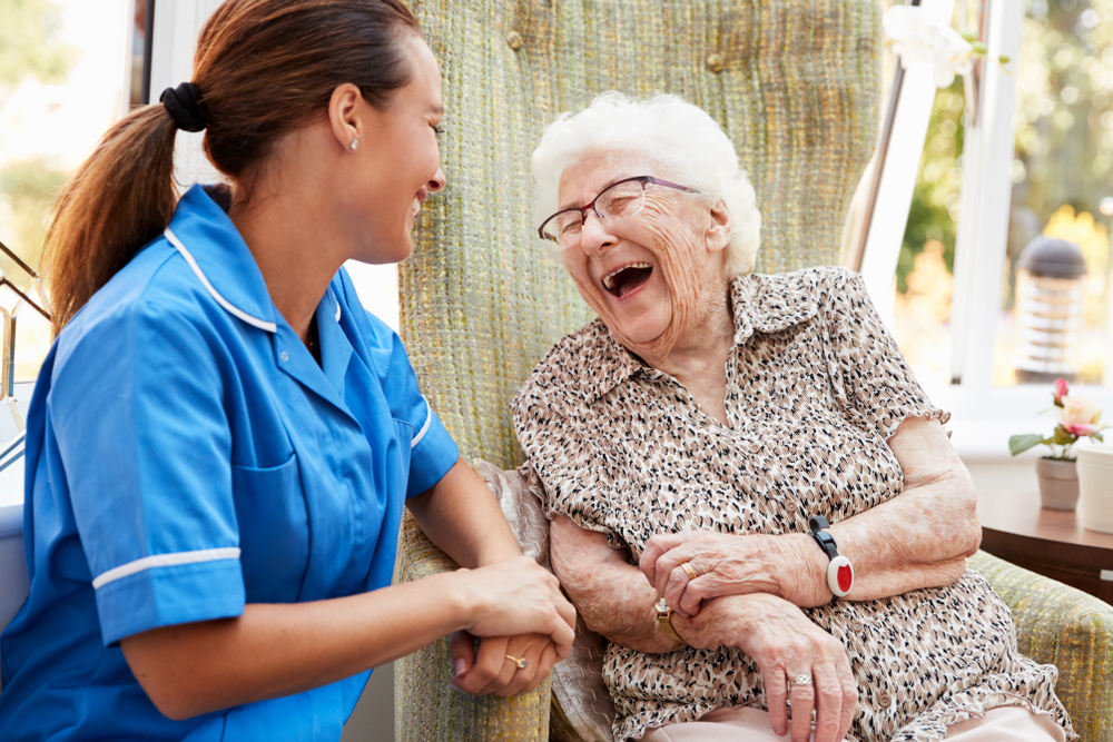 Senior woman sitting in a chair laughing with nurse sitting beside her smiling